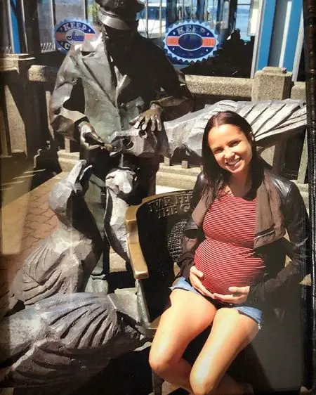 Pregnant Boogie holding her grown belly while sitting on a chair in front of a statue that depicts a man feeding something to and patting an eagle.