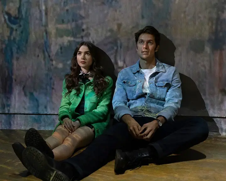 Lily Collins (left) as 'Emily' and Lucas Bravo (right) as 'Gabriel' sitting down leaning against a wall, from 'Emily in Paris'.